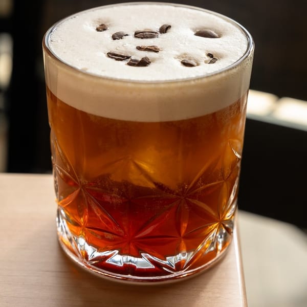 Amber cocktail in a decorative glass, topped with creamy foam and coffee beans, sits on a sunlit wooden table.