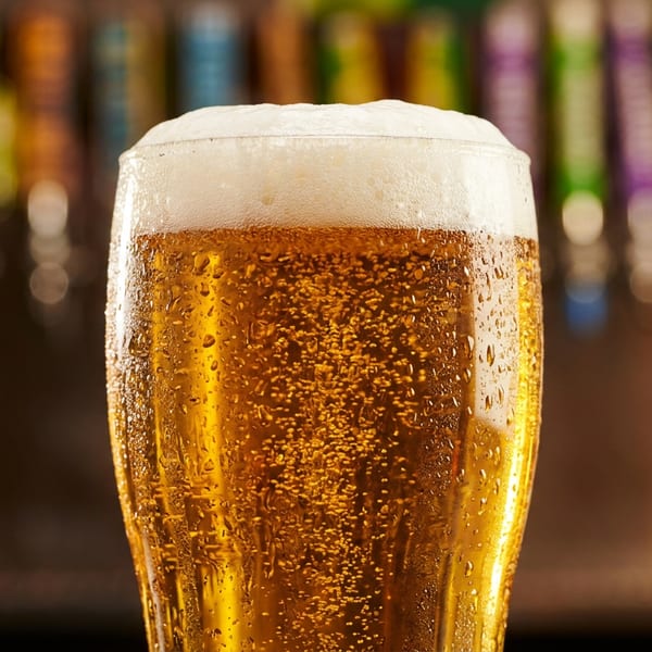 Close-up of a frosty glass of beer with foam and condensation, with blurred beer taps in the background.