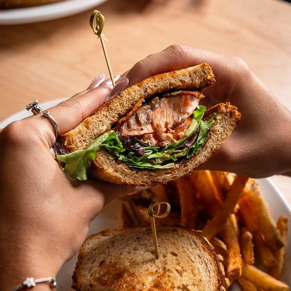 Hands hold a toasted salmon sandwich with leafy greens above a plate of another sandwich and fries.
