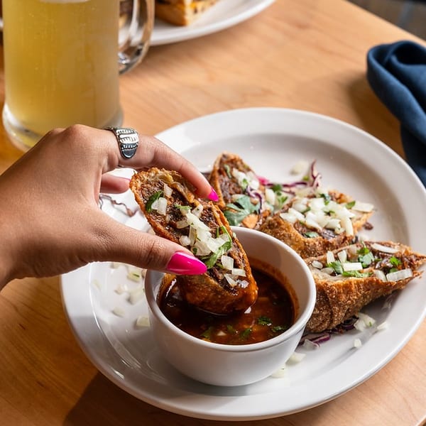 Hand lifts a crispy taco topped with onions and cilantro over consommé, beside more tacos and a beer on a wooden table.