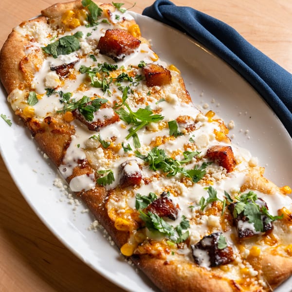 Street corn flatbread topped with crispy pork belly, crema, cotija, and cilantro on an oval plate.