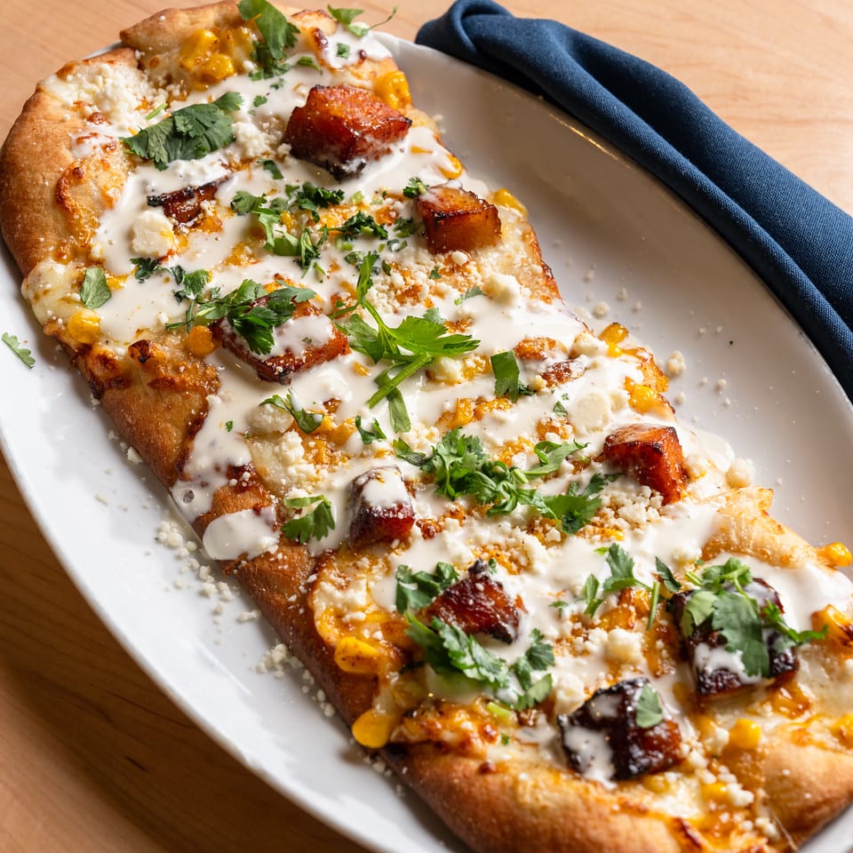 Street corn flatbread topped with crispy pork belly, crema, cotija, and cilantro on an oval plate.