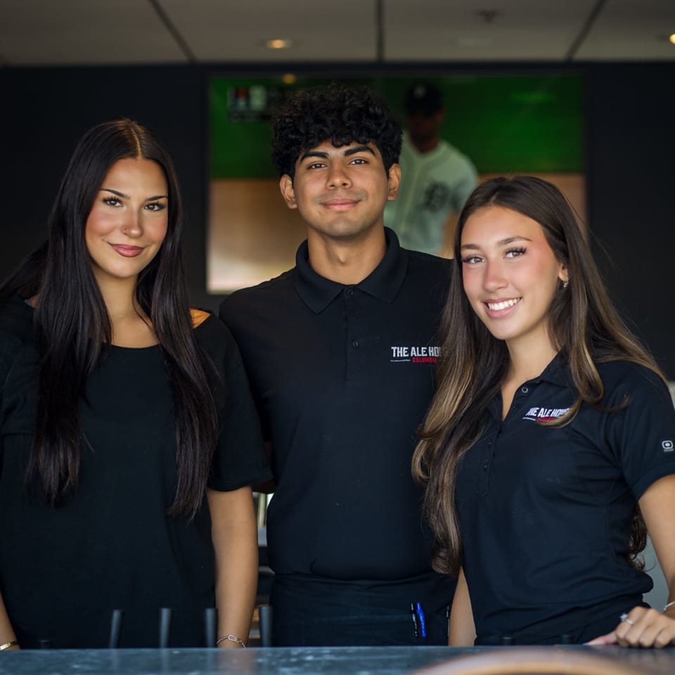 Three smiling Ale House team members behind the bar.