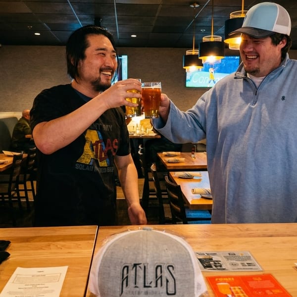 Two men smiling and toasting beers at a bar counter with Atlas branding.