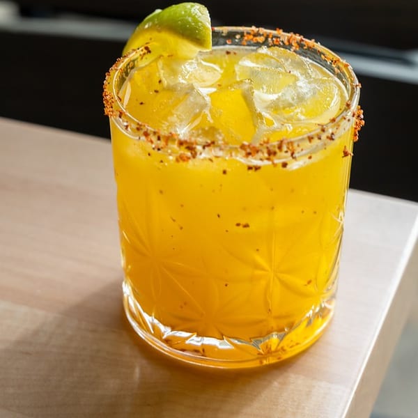 Golden citrus cocktail with ice, chili-salt rim, and lime wedge served in a textured glass on a wooden table.