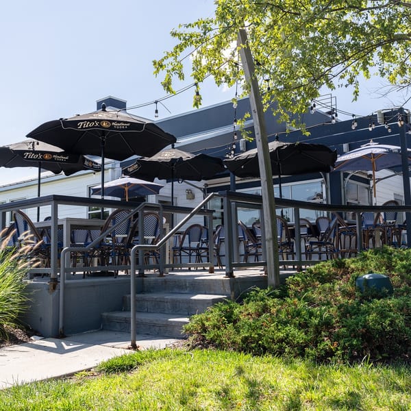 Outdoor restaurant patio with black Tito’s umbrellas, café seating, string lights, stairs, and landscaped greenery.
