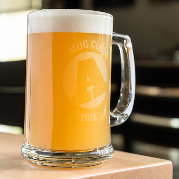 Frosty glass beer mug filled with hazy golden beer, displayed on a wooden bar in a softly lit interior.