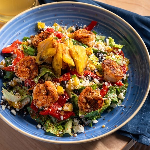 Blue bowl of grilled shrimp salad with roasted vegetables, greens, feta, and crispy plantains beside a drink and napkin.