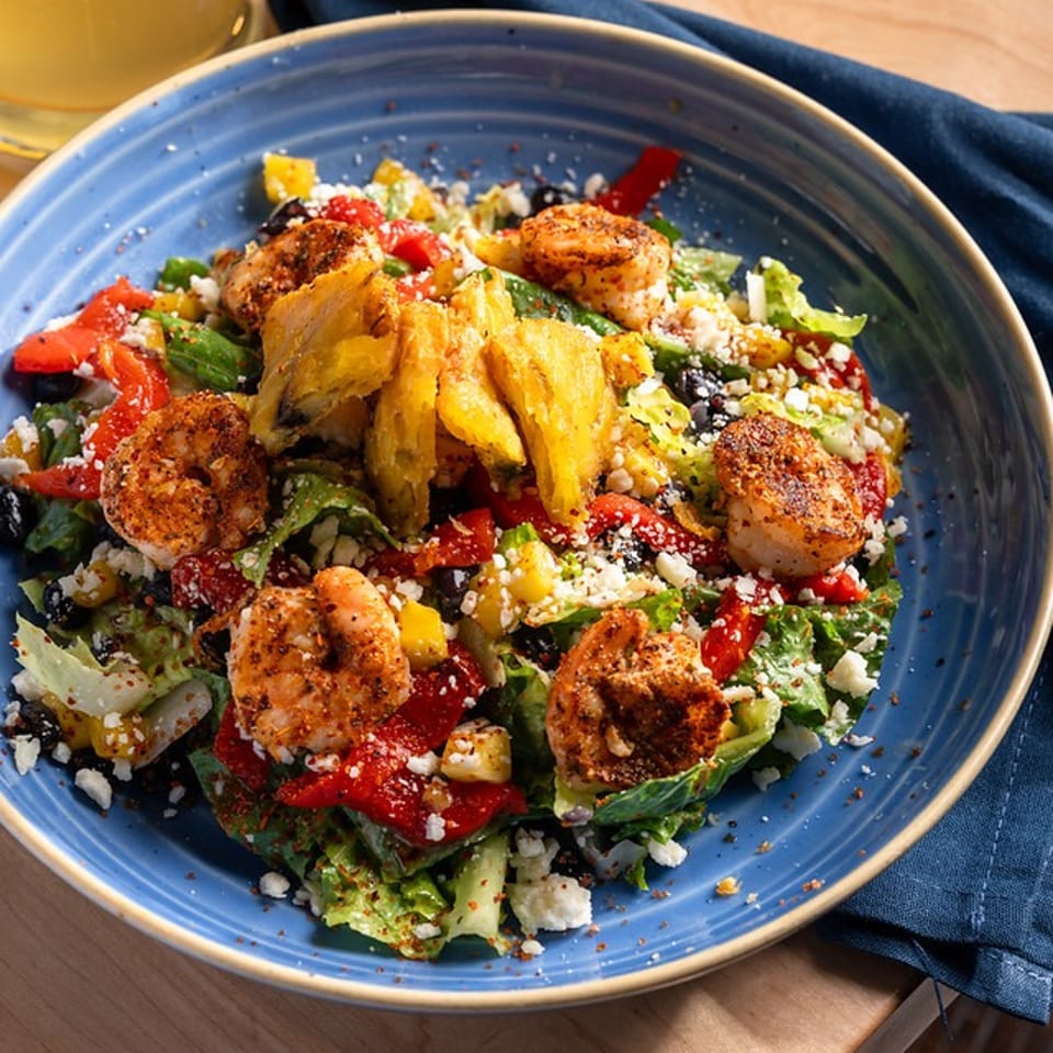 Blue bowl of grilled shrimp salad with roasted vegetables, greens, feta, and crispy plantains beside a drink and napkin.
