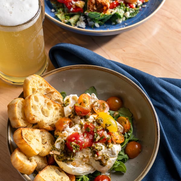 Burrata with cherry tomatoes and toasted baguette beside a shrimp salad and a beer.