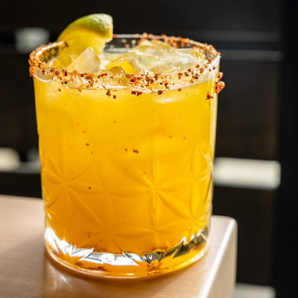 A chili-rimmed mango margarita over ice with a lime wedge on a wooden bar top.