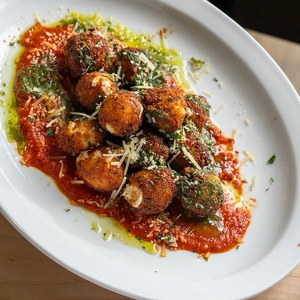 Crispy meatballs topped with grated cheese and herbs, served over tomato sauce and green herb oil on a white plate.